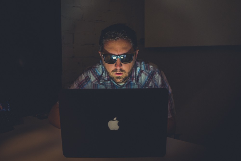 Man som sitter med solglasögon i mörkt rum framför laptop